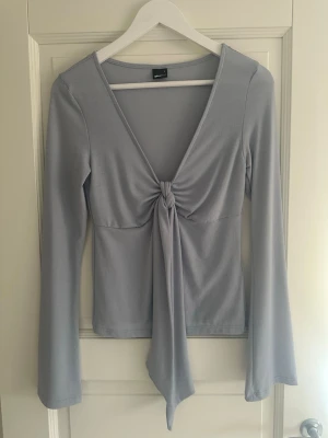 Ljusgrå långärmad topp med knyt - Trendig ljusgrå topp från Gina Tricot med lång ärm och snygg v-ringning. Toppen har ett knyt framtill som ger en cool detalj och är tillverkad i mjukt, stretchigt material. Helt oanvänd!