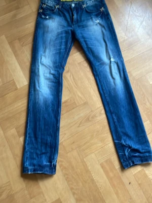 Blå slitna jeans med raka ben  - Säljer ett par blå jeans med slitningar och raka ben från Frankie Morello. Jeansen har klassisk femficksdesign, ljusa tvättade partier och detaljerade sömmar. Perfekta för en avslappnad och trendig look.