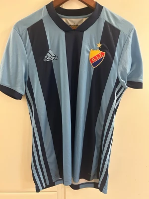 Djurgården matchtröja Adidas blå - Snygg Djurgården fotbollströja från Adidas i ljusblått och marinblått med klubbmärke och stjärna på bröstet. Klassiska Adidas-ränder på sidorna och broderad logga. Perfekt för dig som vill visa ditt stöd på läktaren eller träningen.
