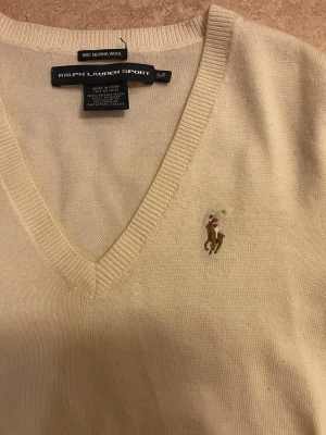 Beige v-ringad tröja från Ralph Lauren - Säljer en beige stickad tröja från Ralph Lauren Sport i 100% merinoull. Tröjan har v-ringning, långa ärmar och ett broderat logotyp på bröstet. Har ett litet hål på framsidan, men syns knappt.