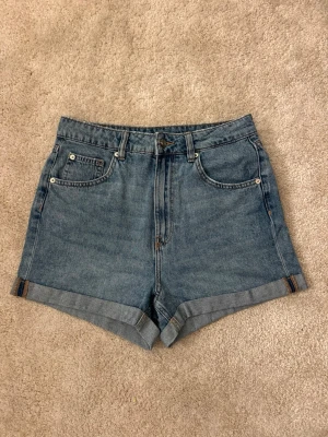 Blå denimshorts med hög midja - Klassiska blå jeansshorts med hög midja och uppvikta benslut. Shortsen har fem fickor och stängs med knapp och dragkedja. 
