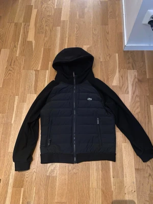 Lacoste cardigan - Snygg svart Cardigan från Lacoste med huva och dragkedja framtill. Jackan har quiltad front och rygg, släta ärmar och två dragkedjefickor. Liten Lacoste-logga på bröstet. Perfekt för kalla dagar och riktigt stilren look.