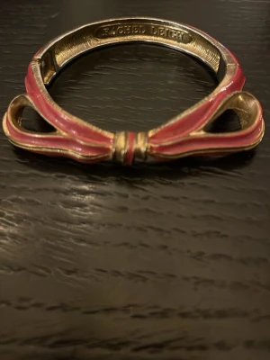 Rachel Leigh armband med rosett - Supersött armband från Rachel Leigh i metall med guldfärgad finish och en lackad rosett i rosa och guld. Armbandet har en unik design där rosetten är i fokus och ger en lekfull men ändå stilren känsla. Perfekt accessoar för att lyfta din outfit.