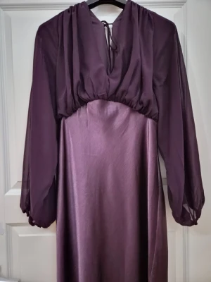 Lila långklänning med v-ringning - Elegant lila långklänning med v-ringning och långa, transparenta ärmar. Klänningen har ett lätt glansigt tyg i kjolen och knytband i nacken. Perfekt för dig som vill ha en stilren och trendig look.