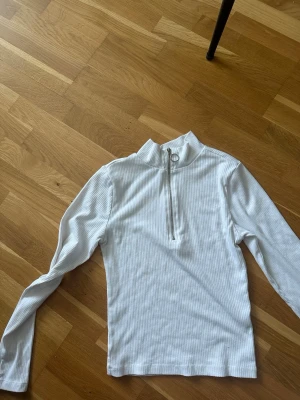 Vit ribbad tröja med dragkedja - Säljer en vit långärmad ribbad tröja med hög krage och silverfärgad dragkedja framtill. Tröjan har en stilren look med diskreta ränder i materialet och en ringdetalj på dragkedjan. Perfekt för en clean och modern stil.