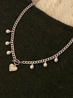 Silverfärgat halsband med pärldroppar - Snyggt silverfärgat halsband med kedja och sju små vita pärldroppar som hänger längs länken. Halsbandet har även ett hjärtformat hänge vid låset som ger en gullig touch. Perfekt accessoar för att lyfta vilken outfit som helst.
