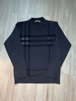 Grå stickad tröja från Burberry - Snygg grå stickad tröja från Burberry med klassiskt rutmönster i mörkgrått och vinrött. Tröjan är Ny i storlek M funkar som S med. Väldigt unik .