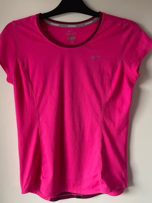 Rosa Nike Dri-Fit löpartröja Storlek M - Snygg rosa löpartröja från Nike med Dri-Fit material som andas. T-shirten har rund halsringning, korta ärmar och en liten Nike-logga på bröstet. Perfekt för träning eller löpning och sitter skönt mot kroppen. Storlek m