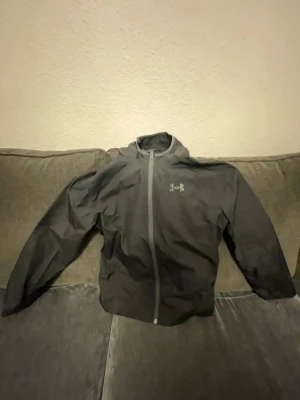 Svart vindjacka från Under Armour - Säljer en svart vindjacka från Under Armour med hel dragkedja och huva. Jackan har ett diskret logotryck på bröstet och meshpanel med stor logga på ryggen för extra ventilation. Perfekt för dig som vill ha en lätt och sportig jacka till träning eller vardag.