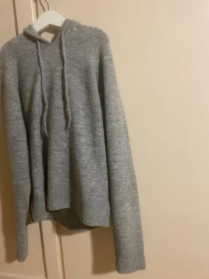 Grå stickad hoodie -  grå stickad hoodie med huva och dragsko. Tröjan har långa ärmar och ribbstickade muddar vid ärmslut och nederkant.