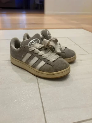 Grå Adidas Campus sneakers - Säljer ett par grå Adidas Campus sneakers i mocka med klassiska vita ränder på sidorna och vit Adidas-logga på plösen och hälen. Skorna har breda vita skosnören och en beige sula som ger en retrovibe. Perfekta för dig som gillar streetwear och enkel stil. ( JAG RENGÖR SKORNA MED MOCKA BORSTE INNAN JAG SICKAR DOM OCH SÅ RENGÖR JAG SLULORNA)🙌🙌🙌🩷🩷