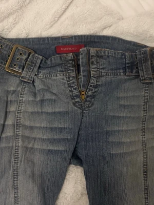Blå bootcut jeans från River Island - Snygga blå bootcut jeans från River Island med slitningar och markerade sömmar. Jeansen har en bred midja med dubbla bälteshällor och medföljande bälte i denim. Klassisk dragkedja och knapp framtill. Innerbenslängden är ca. 82cm pch midjemåttet 34-36 då man kan justera midjan.