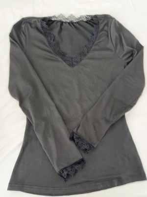 Svart långärmad topp med spetsdetaljer - Stilren svart långärmad topp med v-ringning och dekorativa spetsdetaljer vid halsringning och ärmar.