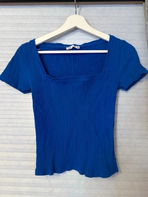 Blå ribbad topp med fyrkantig hals - Snygg blå ribbad topp från PULL&BEAR med korta ärmar och fyrkantig halsringning. Materialet är stretchigt och följsamt, perfekt för att skapa en cool och avslappnad look. Passar dig som gillar stilrena och trendiga plagg.