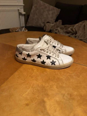 saint laurent skor - Snygga vita sneakers från Saint Laurent med svarta stjärnor längs sidorna. Skorna har klassisk låg profil, vita snören och sula i gummi. Perfekta för dig som vill sticka ut med en cool och unik detalj på fötterna.