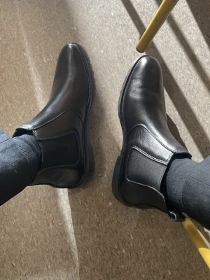 Svarta stövletter i läder - Säljer ett par klassiska svarta stövletter i slätt läder med rund tå och låg klack. Skorna har en enkel och stilren design som passar till många olika outfits. Perfekt för dig som gillar tidlösa boots.