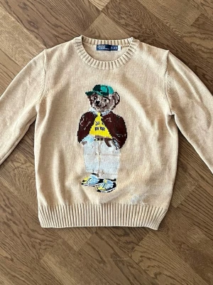 Ralph Lauren beige stickad tröja - Stickad beige tröja från Polo Ralph Lauren med ett stort broderat björnmotiv på framsidan. Björnen har grön keps, brun jacka, gul tröja och sneakers. Tröjan har rund halsringning och ribbade muddar vid ärmslut och nederkant. Tröjan är ganska liten och sitter som en S eller lite mindre. Kolla sista bilderna för mått.