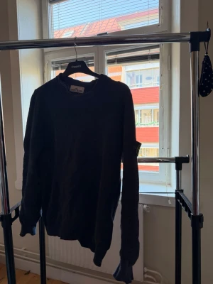 Svart stickad tröja från Stone Island - Säljer en svart stickad tröja från Stone Island med klassisk rund hals och lång ärm. Tröjan har en enkel, stilren design och den ikoniska loggan på vänster ärm. Perfekt för dig som gillar streetwear och vill ha något både snyggt och bekvämt.
