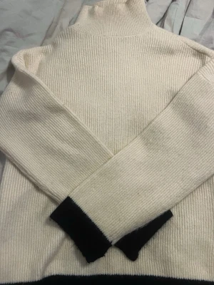 Vit polotröja med svarta detaljer - Stickad polotröja i mjuk, vit ullmix med svarta muddar vid ärmslut och nederkant. Tröjan har ribbstickad struktur och lång ärm, perfekt för kyliga dagar. Enkel och stilren design som passar till det mesta.