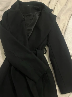 Svart kappa med skärp och bred krage - Stilren svart kappa med bred krage och knytband i midjan. Kappan har släta fickor och ett mjukt, fodrat innerfoder. Perfekt för kyliga dagar och ger en klassisk siluett. Materialet ser ut att vara ullblandning.