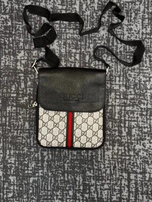 axelväska med GG-mönster - Snygg axelväska med klassiskt GG-mönster i beige och svart. Väskan har svart lock i läder, justerbar axelrem och en ikonisk röd-grön rand framtill. Perfekt för dig som vill ha en trendig och stilren accessoar.