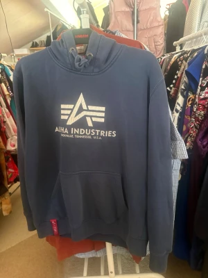 Blå hoodie från Alpha Industries - Snygg blå hoodie från Alpha Industries med vit logga och text på bröstet. Klassisk känguruficka framtill, ribbade muddar och justerbar huva med dragsko. Perfekt för en avslappnad och cool stil.