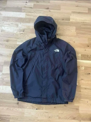 Svart vindjacka från The North Face - Svart vindjacka från The North Face med huva och dragkedja. Jackan har två snedställda fickor framtill och elastiska muddar vid ärmslut och midja. Perfekt för blåsiga dagar och enkel att matcha med olika outfits.