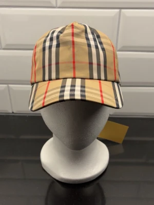 Beige keps med rutigt mönster Burberry - Snygg beige keps från Burberry med klassiskt rutigt mönster i svart, vitt och rött. Kepsen har böjd skärm och justerbar rem baktill. Perfekt accessoar för en trendig och stilren look.