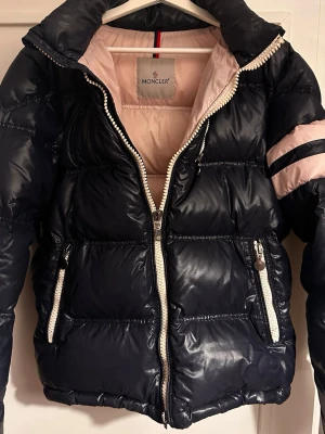 Blå dunjacka från Moncler - Snygg blå dunjacka från Moncler. Jackan har två fickor med dragkedja och Moncler-logga på vänster ärm. 