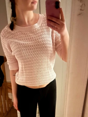Ljusrosa stickad kortärmad tröja - Säljer en ljusrosa stickad tröja med korta ärmar och rund hals. Tröjan har ett luftigt, hålmönstrat stickat material och ribbade muddar vid ärmslut och hals. Perfekt för lager-på-lager och en chill stil.