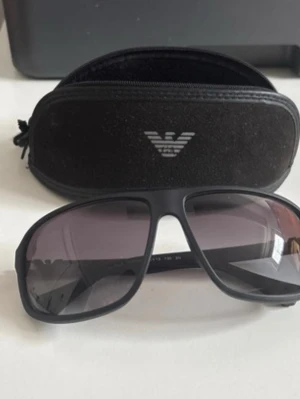 Svarta solglasögon från Emporio Armani - Snygga svarta solglasögon från Emporio Armani med klassisk pilotform. Bågarna är i svart plast med metalliga detaljer på sidorna och logga på skalmarna. Kommer med originalfodral. Perfekta för dig som vill ha en stilren och modern look.