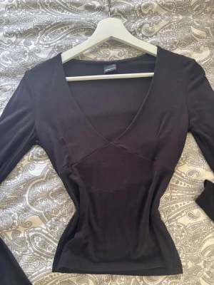 Svart v-ringad topp från Gina Tricot - Snygg svart långärmad topp från Gina Tricot med v-ringning och figurnära passform. Perfekt att styla med jeans eller kjol för en trendig look. Tillverkad i mjukt material som känns skönt mot huden.