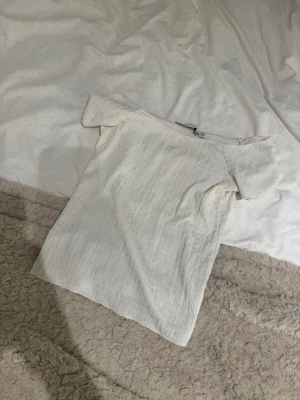 Vit ribbad offshoulder topp - Snygg vit topp med ribbad struktur och offshoulder-design. Perfekt att styla med jeans eller kjol för en trendig look. Tunt och mjukt material som känns skönt mot huden.