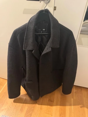 Mörkgrå ullkappa från 157 - Stilren mörkgrå kappa från 157 med klassisk krage och stora fickor framtill. Kappan är tillverkad i ett tjockt ullmaterial och har en rak passform. Perfekt för kyliga dagar och ger en tidlös look.