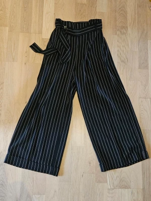 Svarta vida randiga kostymbyxor, Inwear - Säljer ett par svarta kostymbyxor med vvertikala ränder och hög midja. Från Inwear, modell Jacey Culotte pants. Byxorna har ett brett ben, vida benslut och ett matchande bälte i midjan. Perfekta för en trendig och avslappnad look. Aldrig använda. Nypris 1300kr