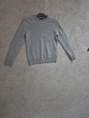 Grå stickad tröja från Gant - Klassisk grå stickad tröja från Gant med rund halsringning och diskret logga på bröstet. Tröjan har långa ärmar och ribbade muddar vid ärmslut och nederkant. Perfekt för lager-på-lager och enkel att matcha.