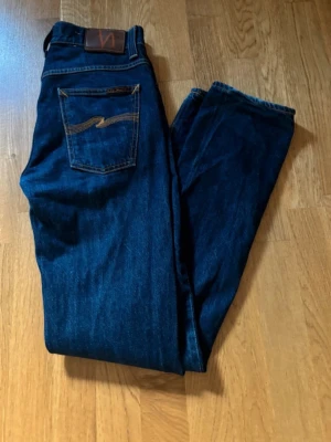 Mörkblå jeans från Nudie Jeans - Snygga mörkblå jeans från Nudie Jeans med klassisk rak passform och femficksdesign. Jeansen har tydliga kontrastsömmar och en ikonisk broderad bakficka. Tillverkade i slitstark denim av bomull.