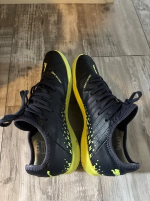 Puma svarta och gula inomhus fotbollsskor - Säljer ett par snygga fotbollsskor från Puma i svart med gula detaljer och sula. Skorna har snörning, låg profil och coolt blixtmönster på sidorna. Perfekta för dig som vill sticka ut på planen och få bra grepp och kontroll.