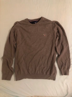 Grå stickad tröja från GANT - Säljer en brun stickad tröja från GANT med rund halsringning och diskret logga broderad på bröstet. Tröjan har långa ärmar och ribbade muddar vid ärmslut och nederkant. Perfekt för lager-på-lager och enkel att matcha.