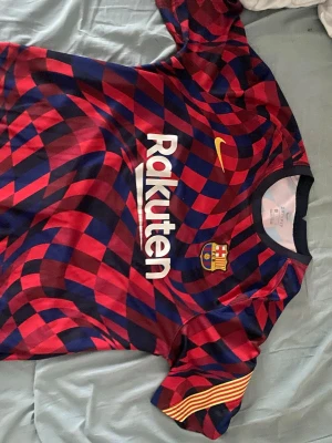 Barcelona  Nike röd/blå - Säljer en Barcelona matchtröja från Nike med ett unikt röd- och blårutigt mönster. Tröjan har klubbmärket, Rakuten och Beko-loggor samt gula detaljer på ärmen. Tillverkad i lätt och ventilerande polyester, perfekt för fotboll eller träning.
