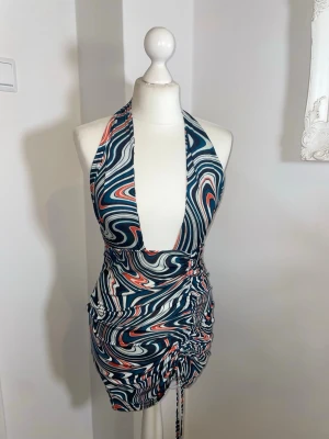 Mönstrad halterneck miniklänning - Säljer en snygg miniklänning med halterneck och djup urringning. Klänningen har ett färgstarkt swirl-mönster i blått, vitt och orange samt rynkade detaljer med dragsko på sidan. Materialet är stretchigt och följsamt, perfekt för dig som gillar statement-plagg.