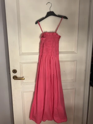 Rosa långklänning med smock - Säljer en rosa långklänning med smock upptill och utställd kjol. Klänningen har en enkel och stilren design, perfekt för varma dagar. Materialet känns lätt och luftigt, vilket gör den skön att bära. Använt två gånger och finns inga tydliga tecken på användning.