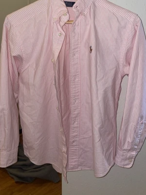 Rosa och vit randig skjorta Polo Ralph Lauren - Säljer en klassisk rosa och vit randig skjorta från Polo Ralph Lauren med broderad logga på bröstet. Skjortan har lång ärm, knappar framtill och button-down krage. Tillverkad i mjuk bomull och passar perfekt till jeans eller chinos.