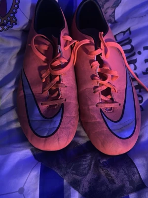Två par Nike fotbollsskor - Säljer två par Nike fotbollsskor. Ett par är orange med svart swoosh och snörning, det andra paret är gult med limegröna detaljer och svart swoosh. Båda paren har låg profil och är tillverkade i syntetmaterial för bra bollkontroll och grepp på planen.