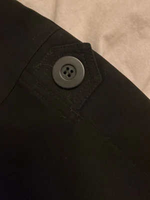 Svarta kostymbyxor med knappdetalj - Säljer ett par svarta kostymbyxor med snygg knappdetalj vid nedre benet. Byxorna har rak passform och är tillverkade i ett slätt tyg som ger en clean look. Perfekta för dig som vill ha en stilren och enkel byxa till garderoben.