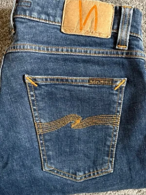 Blå jeans från Nudie Jeans - Säljer ett par klassiska blå jeans från Nudie Jeans med ikonisk söm på bakfickan och läderpatch i midjan. Jeansen har fem fickor och är tillverkade i mjuk denim med stretch för extra komfort. Perfekta för en avslappnad och trendig look.