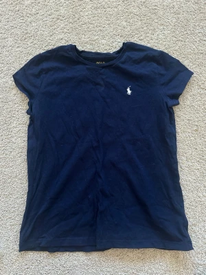 Marinblå t-shirt från Polo Ralph Lauren - Klassisk marinblå t-shirt från Polo Ralph Lauren med korta ärmar och rund hals. T-shirten har den ikoniska vita logon broderad på bröstet och är tillverkad i mjuk bomull. Perfekt basplagg till garderoben.