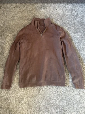 Brun halvzip-tröja från Massimo Dutti - Snygg brun långärmad tröja från Massimo Dutti med halv dragkedja och hög krage. Tillverkad i bomull för en skön och avslappnad känsla. Perfekt till jeans eller chinos för en clean look.