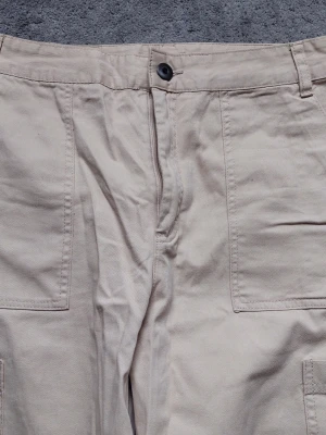 Beiga cargobyxor med raka ben - Säljer ett par beiga cargobyxor med raka ben och stora fickor på sidorna. Byxorna har hög midja, bälteshällor och stängs med knapp och dragkedja. Materialet känns slitstarkt och passar perfekt för en avslappnad streetwear-look.