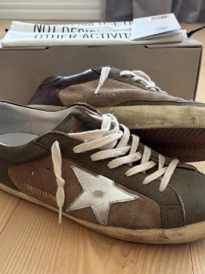 Golden Goose Superstar - Golden Goose Superstar sneakers i olivgrönt och brunt mocka med vit stjärndetalj på sidan och vita skosnören. Klassisk låg modell med slitna detaljer och vit sula. Riktigt bra skick, skriv vid minsta fråga eller fundering! 